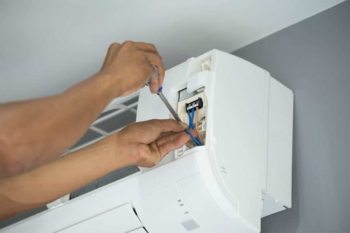 A close-up, horizontal image showing two hands, one holding a screwdriver and the other holding a blue wire, performing maintenance on the electrical components inside a wall-mounted white air conditioning unit. The unit's cover is partially open.