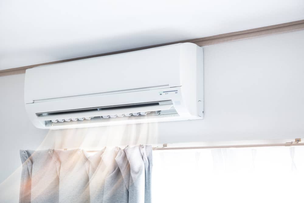 A wall-mounted white split-system air conditioner blowing air onto light grey curtains.