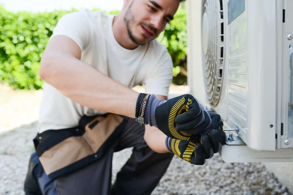 A focused technician wearing protective gloves is seen tightening a screw on an outdoor air conditioning unit.