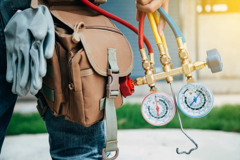 A close-up shot of an HVAC technician holding a refrigerant manifold gauge set with red, yellow, and blue hoses. The technician is wearing a tool belt and gloves.