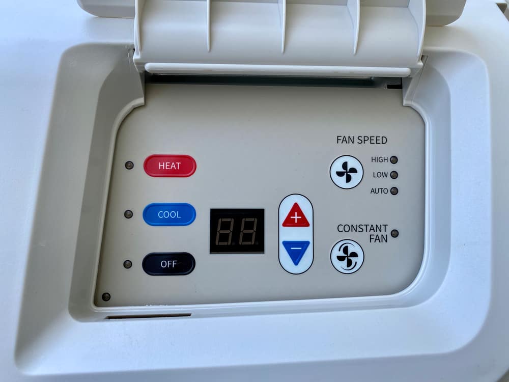 Close-up of a PTAC Air Conditioning & Heating NYC control panel on a white appliance, featuring buttons for Heat (red), Cool (blue), Off (black), fan speed options, constant fan, and temperature adjustment.