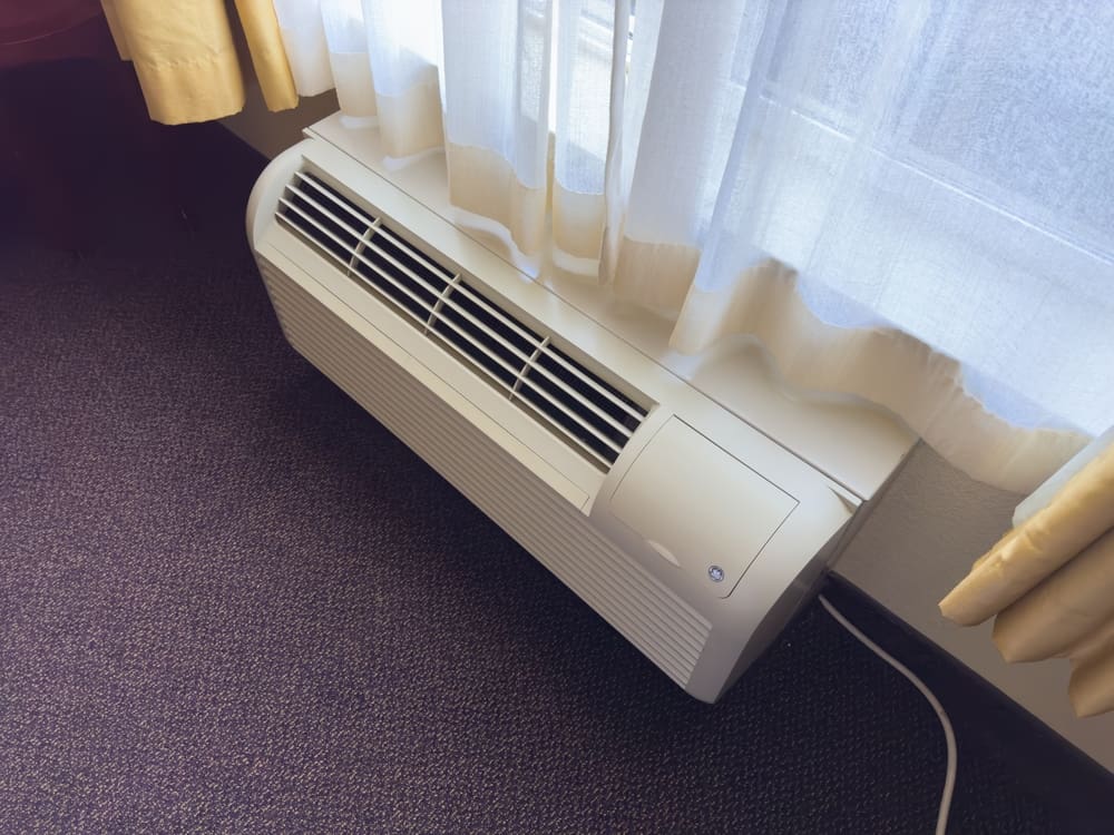 An off-white packaged terminal air conditioner (PTAC) unit is shown from a high angle, situated on a dark carpeted floor below a window with white and yellow curtains.
