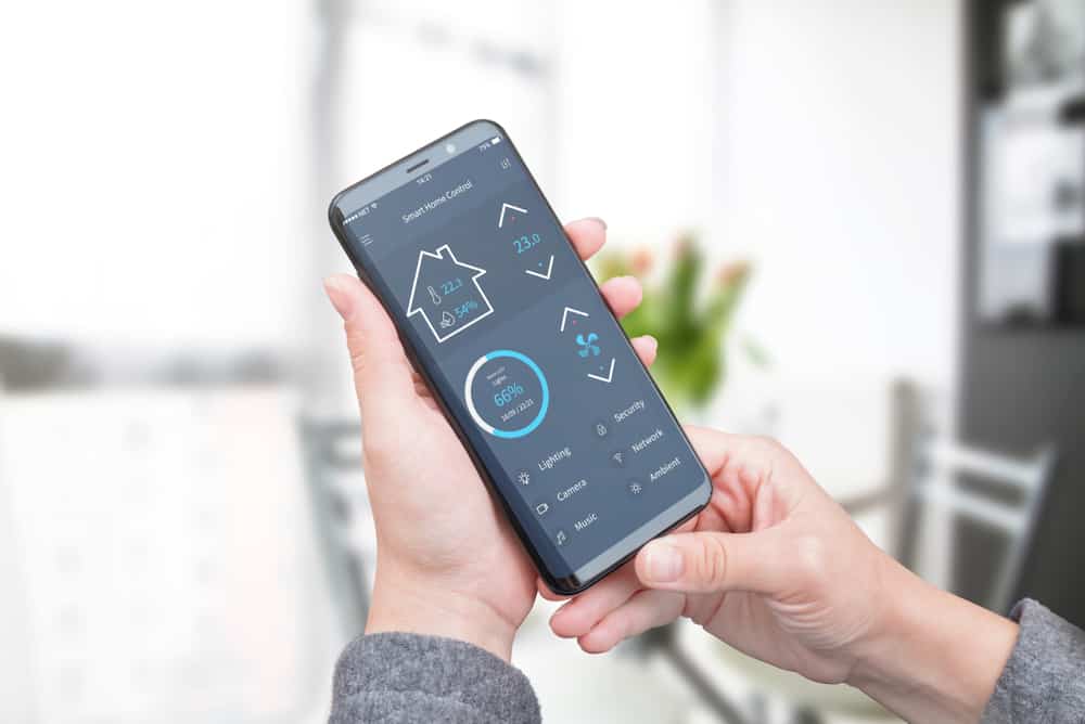 Hands holding a smartphone displaying a smart home control app with a dashboard for a house's temperature, lighting, and security.