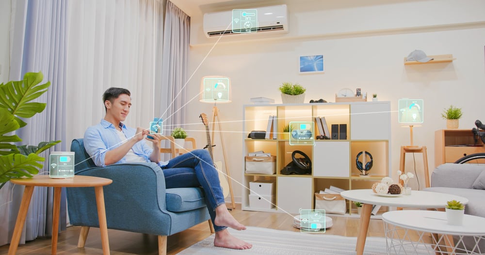 A man sitting in a living room using his smartphone to control various smart home devices represented by glowing digital icons and connecting lines.