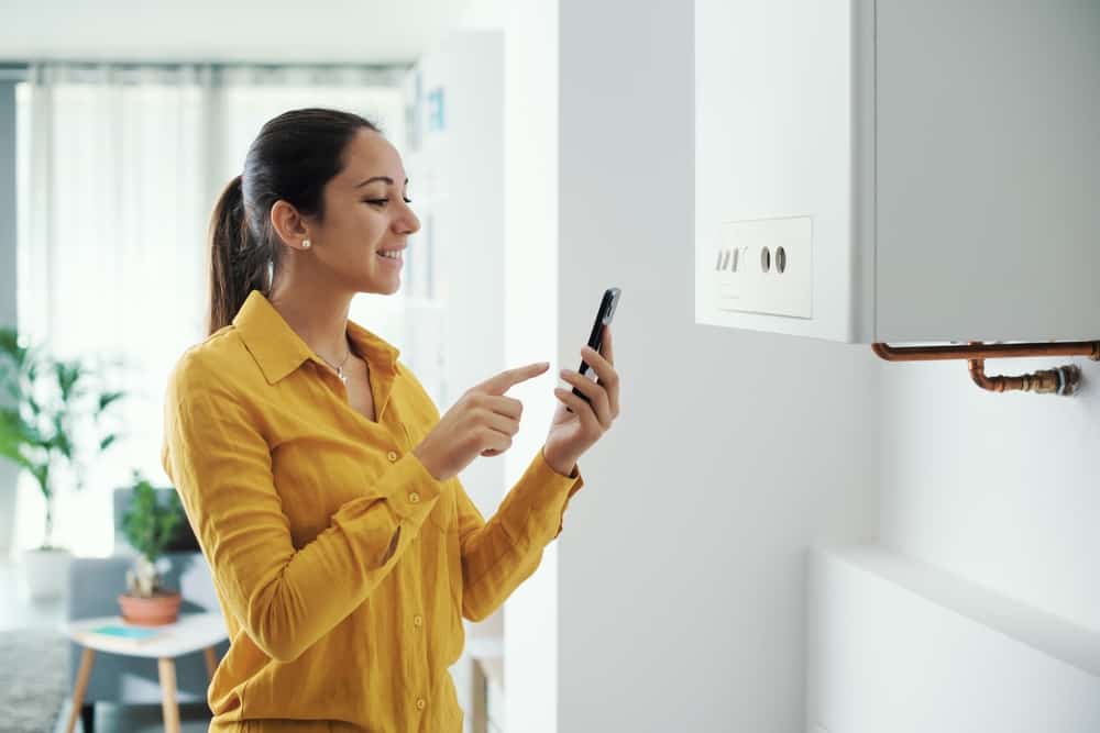 A smiling woman using her smartphone to control a wall-mounted boiler or HVAC unit.