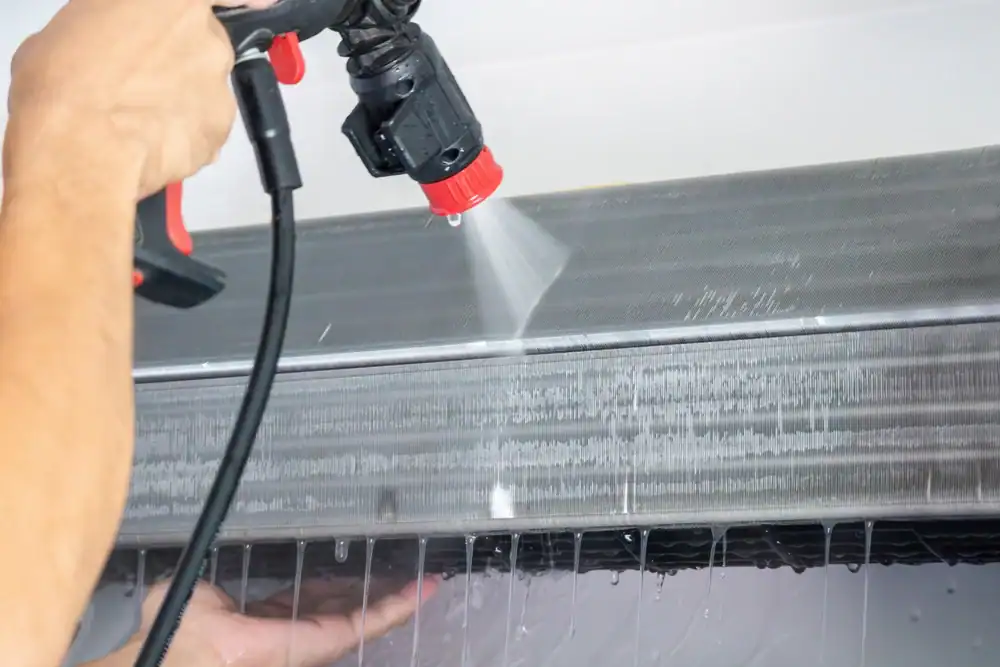 A person sprays water to clean the interior filter of a PTAC Air Conditioning & Heating NYC unit, with water droplets visible dripping down from the appliance.