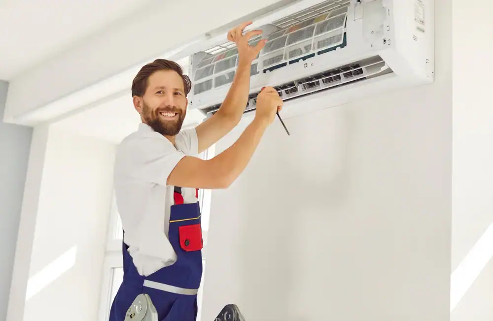 A smiling technician in overalls stands on a ladder while installing or repairing a wall-mounted air conditioner using a screwdriver, representing PTAC Air Conditioning & Heating NYC’s expert service.
