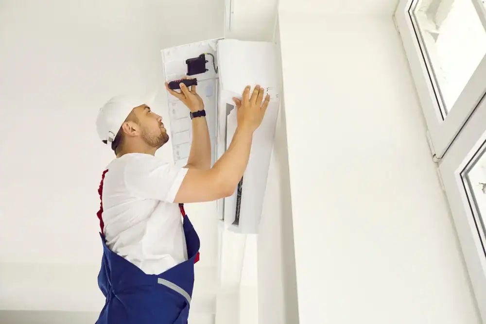 A technician in a white shirt, blue overalls, and a white cap repairs or installs a wall-mounted PTAC Air Conditioning & Heating NYC unit near a large window.
