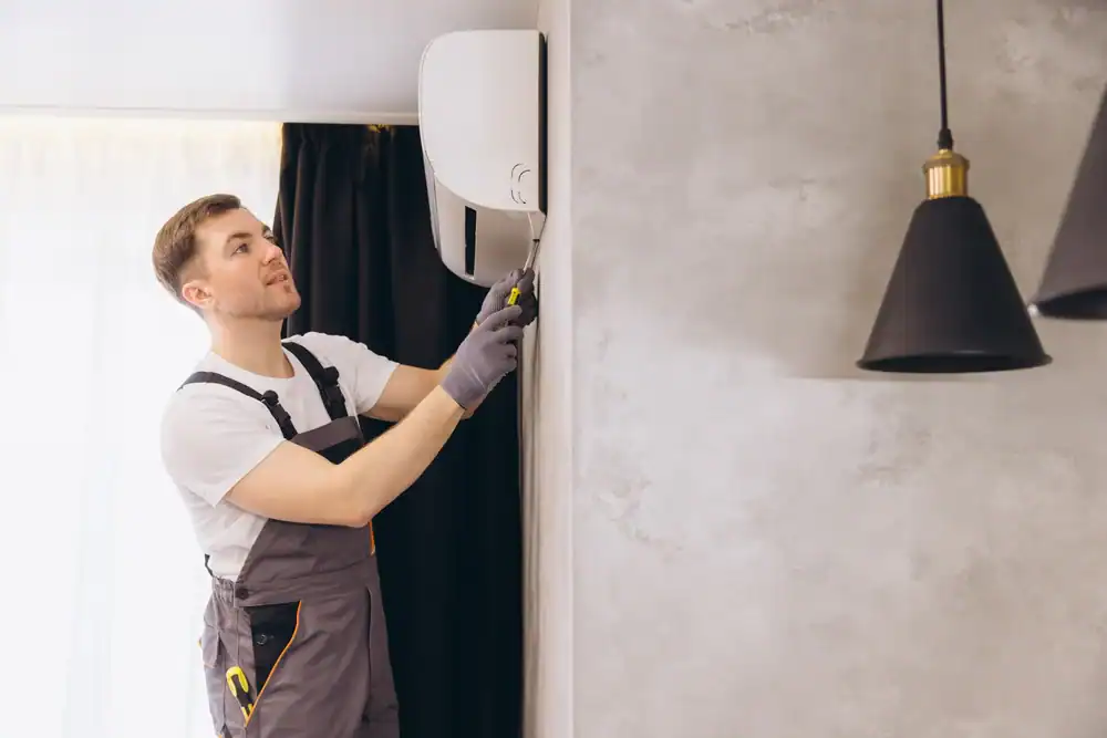 A repairman wearing overalls and gloves is using a screwdriver to fix or install a wall-mounted PTAC Air Conditioning & Heating NYC unit in a modern room with grey walls and black pendant lights.
