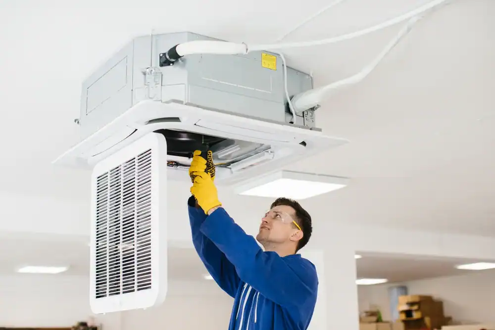 A technician wearing safety glasses and yellow gloves is servicing or installing a ceiling-mounted PTAC Air Conditioning & Heating NYC unit in a bright indoor setting.