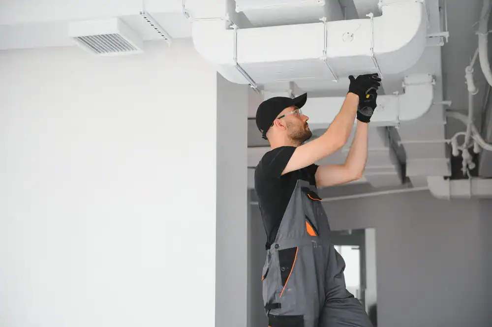A worker in overalls and a cap is fixing or inspecting an air duct on the ceiling inside a modern building. He is using tools and wearing gloves for safety, ensuring top-quality PTAC Air Conditioning & Heating NYC system performance.