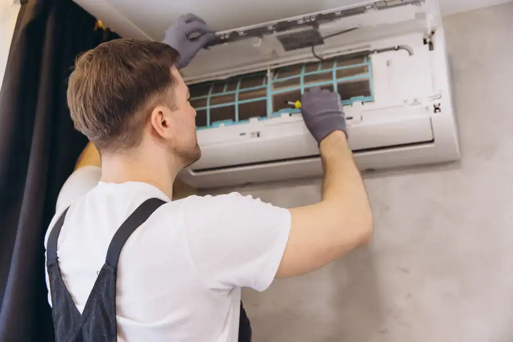 A person wearing gloves and overalls is servicing a wall-mounted air conditioner, with the front panel open and tools in hand—typical of expert PTAC Air Conditioning & Heating NYC maintenance.