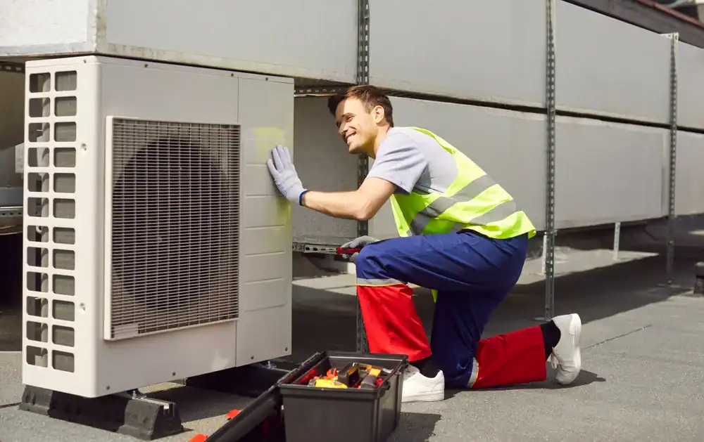 A worker in a reflective vest and gloves kneels beside a large outdoor HVAC unit, inspecting it. A PTAC Air Conditioning & Heating NYC toolbox with various tools is open on the ground nearby.