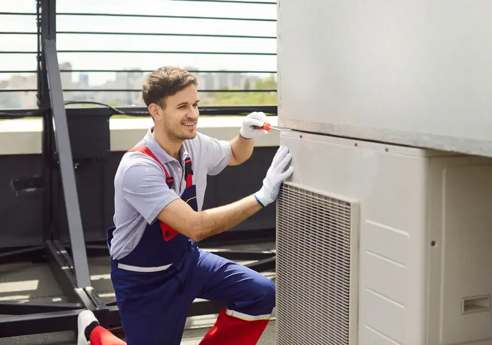 A male technician in overalls and gloves is kneeling while working on a PTAC Air Conditioning & Heating NYC unit on a rooftop, using a screwdriver and smiling.