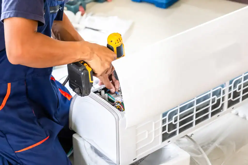 A technician in overalls uses a power drill to perform emergency HVAC repair on a wall-mounted air conditioning unit, with tools and wiring visible inside the open panel.