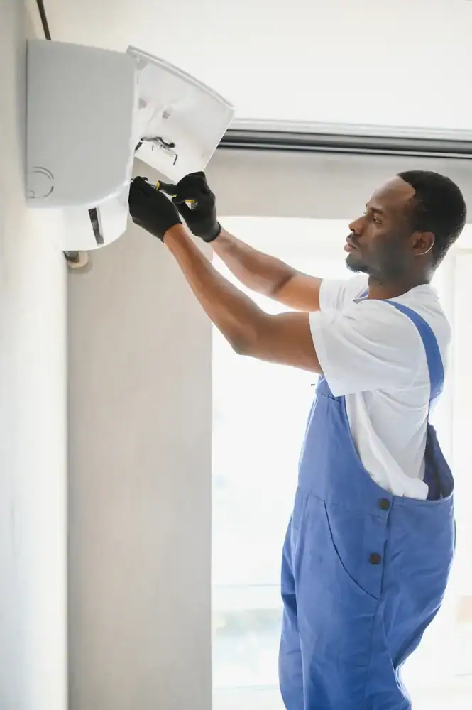 A technician wearing blue overalls and black gloves repairs or installs a PTAC Air Conditioning & Heating NYC unit mounted on a wall.