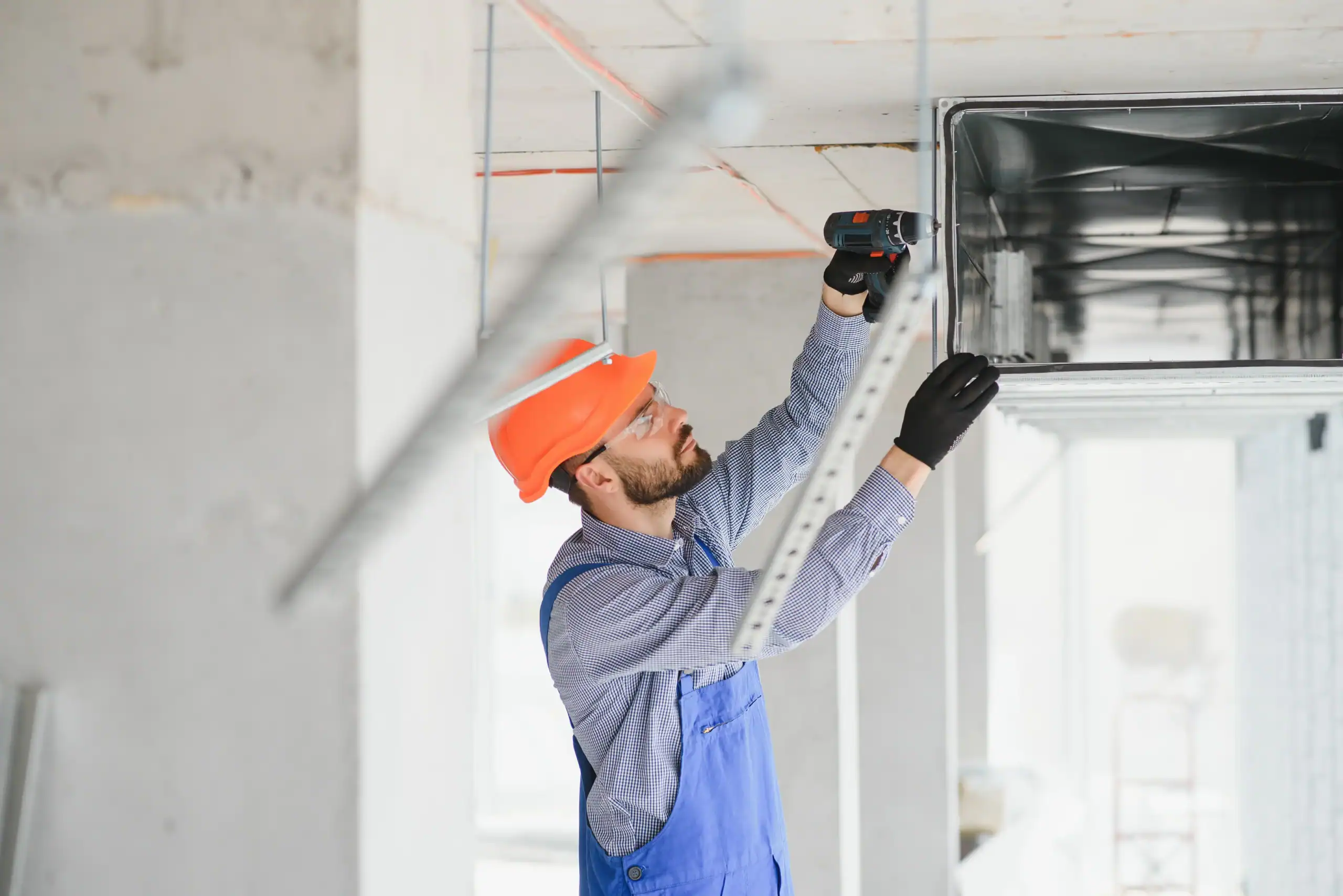 A construction worker wearing a safety helmet and gloves installs or repairs an overhead ventilation duct using a power drill inside an unfinished building for PTAC Air Conditioning & Heating NYC.